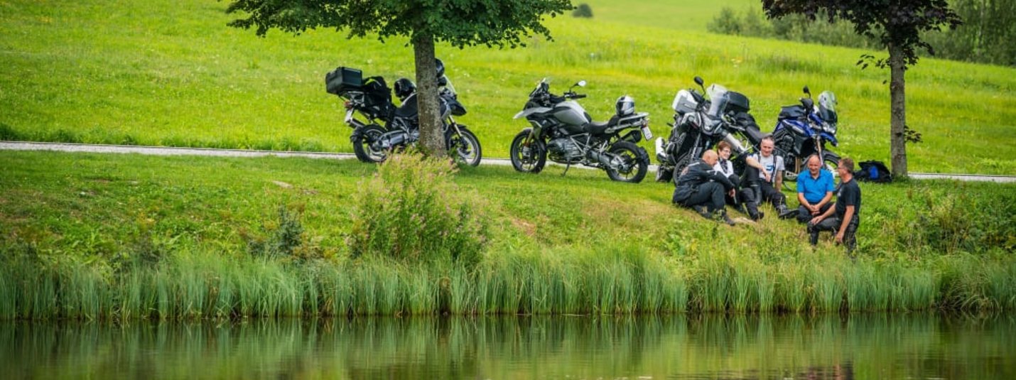 MOTORRAD - Genuss-Tage So-Do 2026 Gruppe von Motorradfahrern entspannt sich an einem See in grüner Landschaft