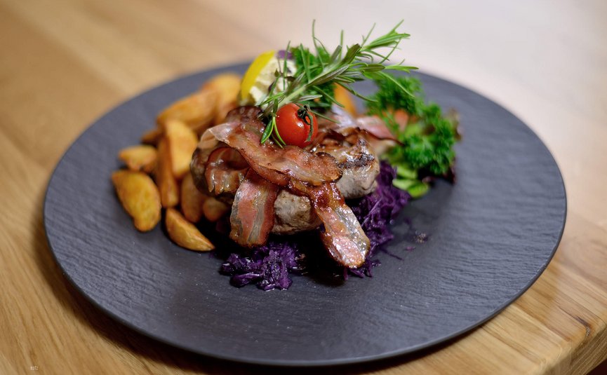 Gegrilltes Fleisch mit Speck, Kartoffelspalten und Kräutern auf schwarzem Teller