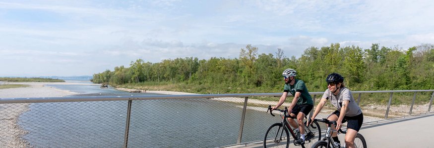 Zwei Radfahrer auf einer Brücke neben einem Fluss unter bewölktem Himmel