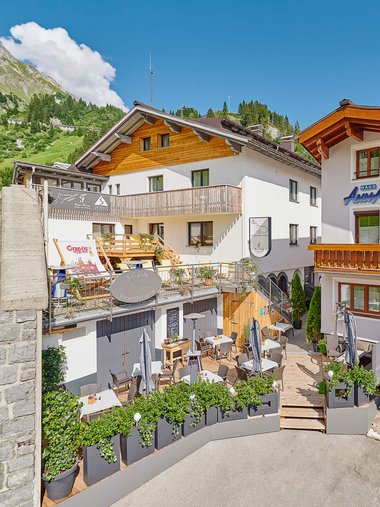 Gastgarten mit Tischen und Stühlen vor Häusern in Alpenlandschaft
