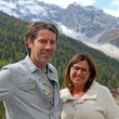 Ein Mann und eine Frau vor bewaldeten Bergen und schneebedecktem Gipfel