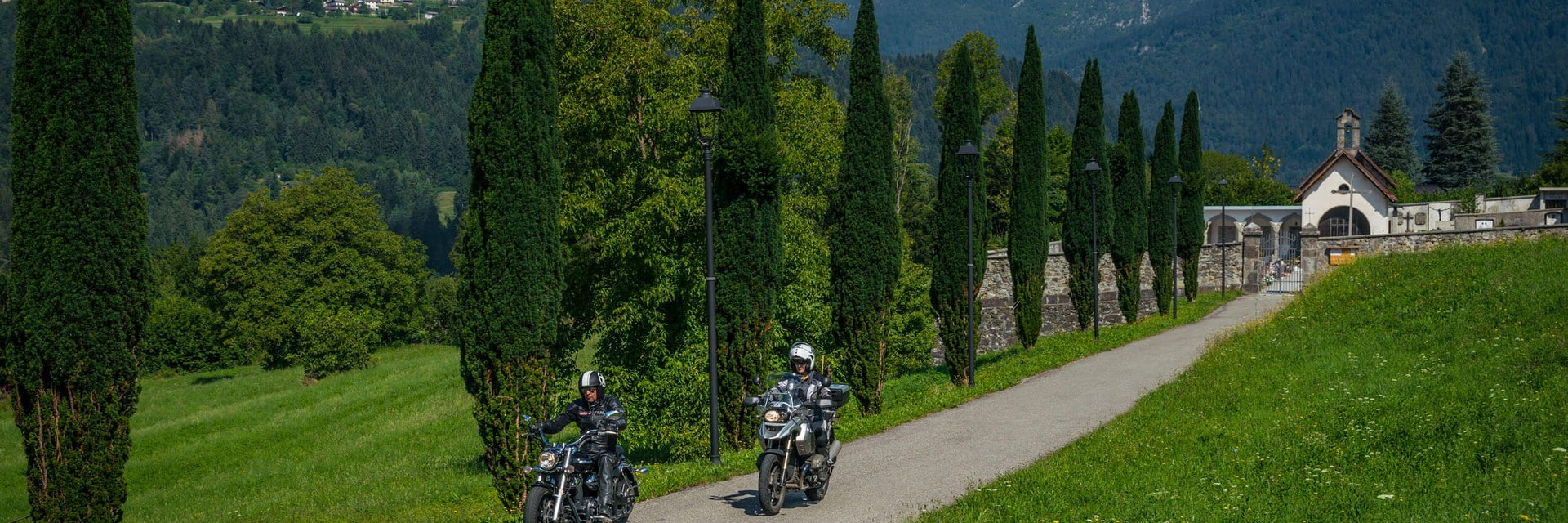 Friaul – Karnien © Moppetfoto.de Zwei Motorradfahrer fahren auf einem Weg mit Bäumen und Bergen im Hintergrund
