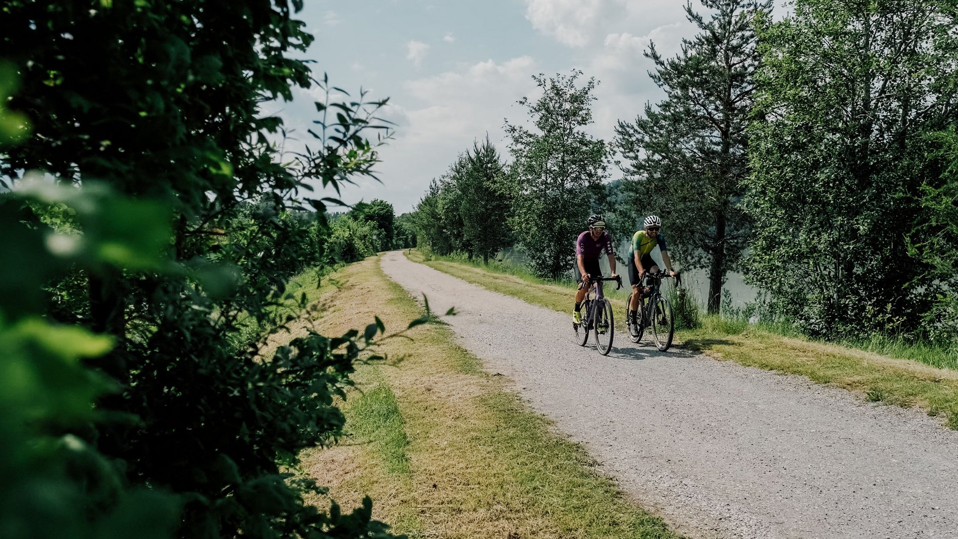 Gravel-Wundertüte Wörthersee © Region Wörthersee Rosental Tourismus GmbH - Chris Perkles Zwei Radfahrer auf einem Waldweg neben einem See unter bewölktem Himmel
