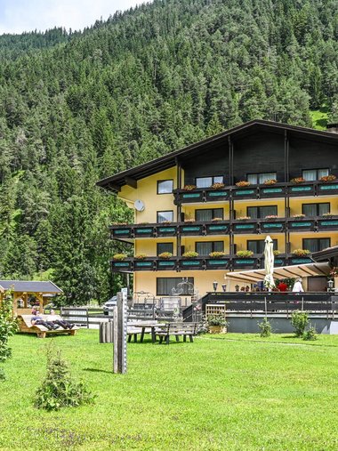 Gelbes Alpenhotel mit Garten vor bewaldetem Berg im Sommer
