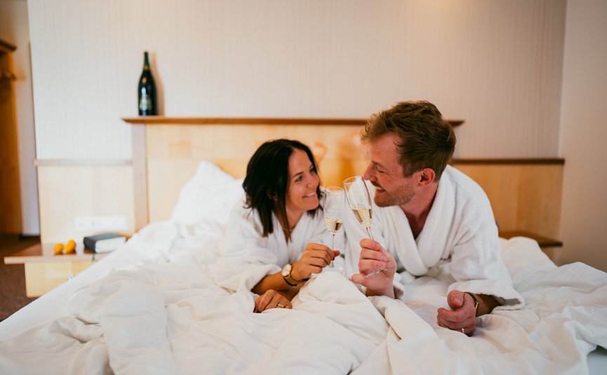 Paar in Bademänteln auf Bett, stößt mit Sektgläsern an