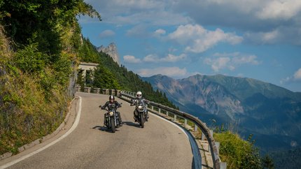 Zwei Motorradfahrer auf kurviger Bergstraße mit Bergen im Hintergrund.