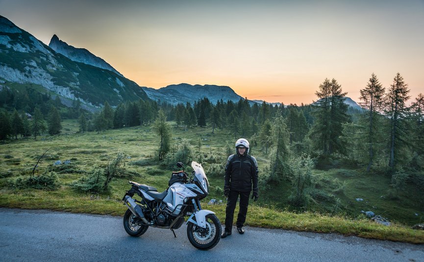 Motorradfahrer mit Helm neben Motorrad vor Berglandschaft bei Sonnenaufgang