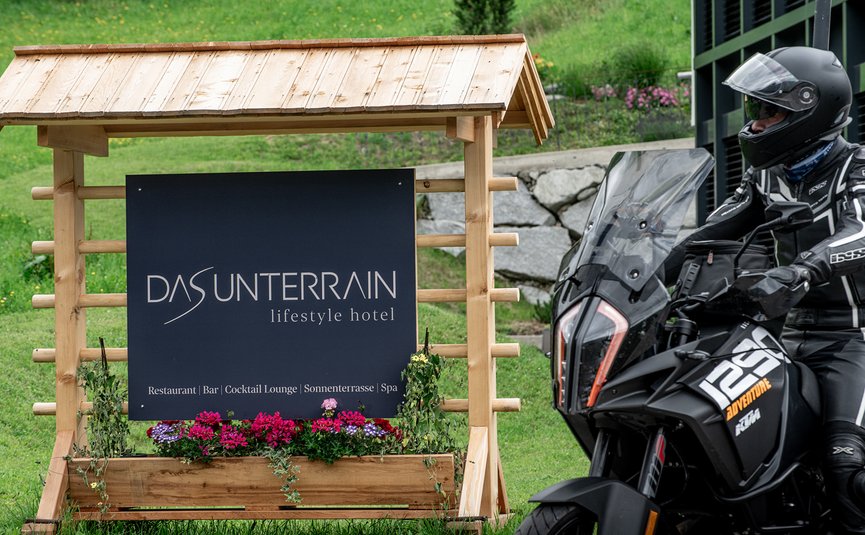 DAS UNTERRAIN lifestyle hotel **** © Armin Kleinlercher Motorradfahrer neben Holzschild vom Hotel Das Unterrain mit Blumen