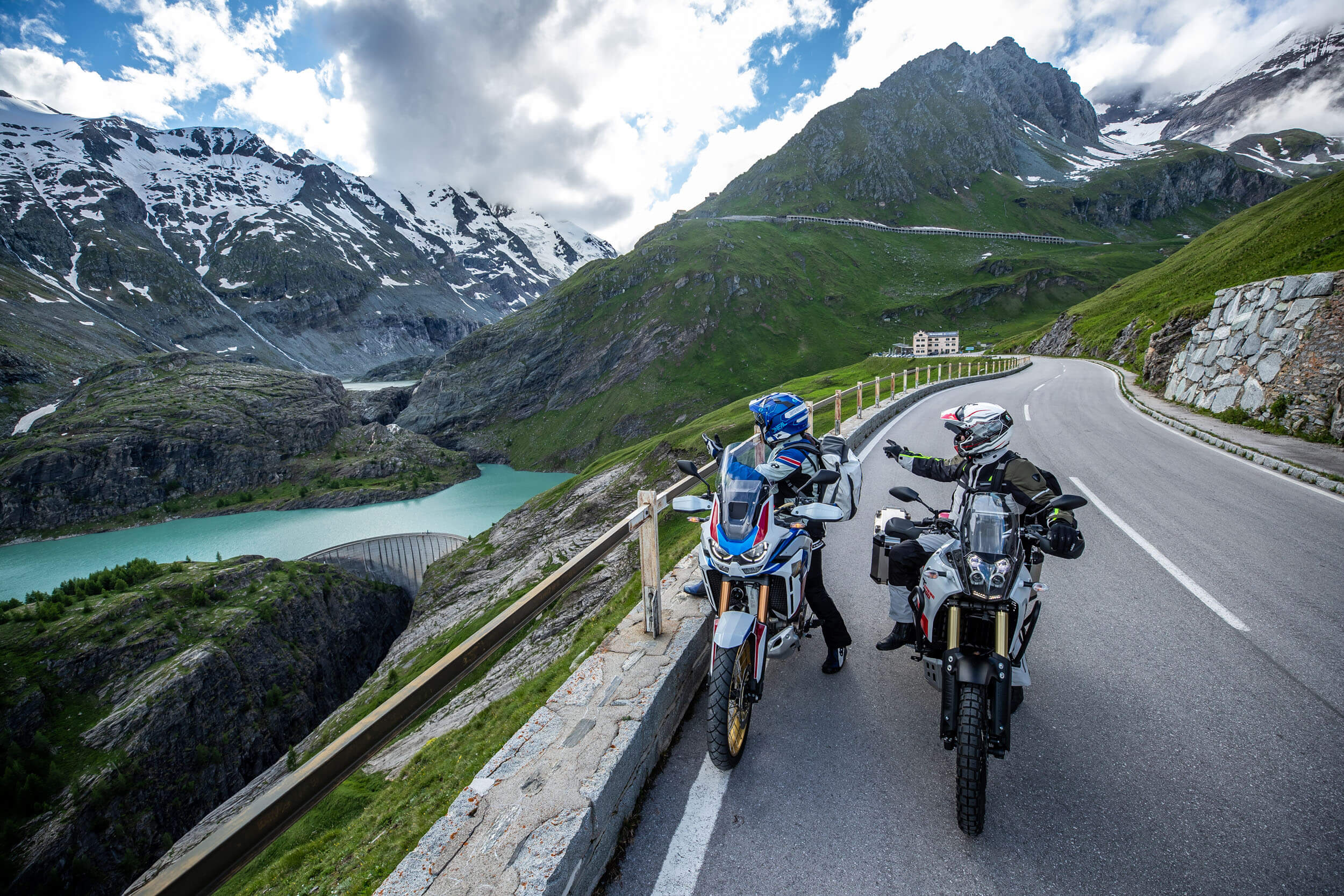 Zwei Motorradfahrer auf Alpenstraße mit Bergsee und schneebedeckten Gipfeln