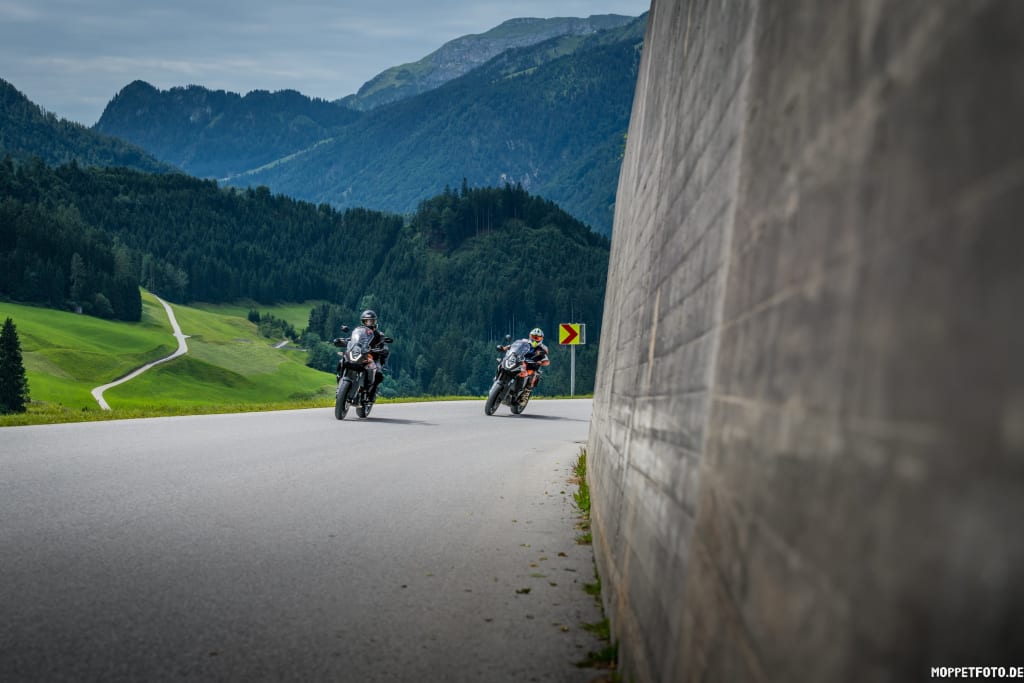 Zwei Motorradfahrer auf Bergstraße mit Wald und Bergen im Hintergrund