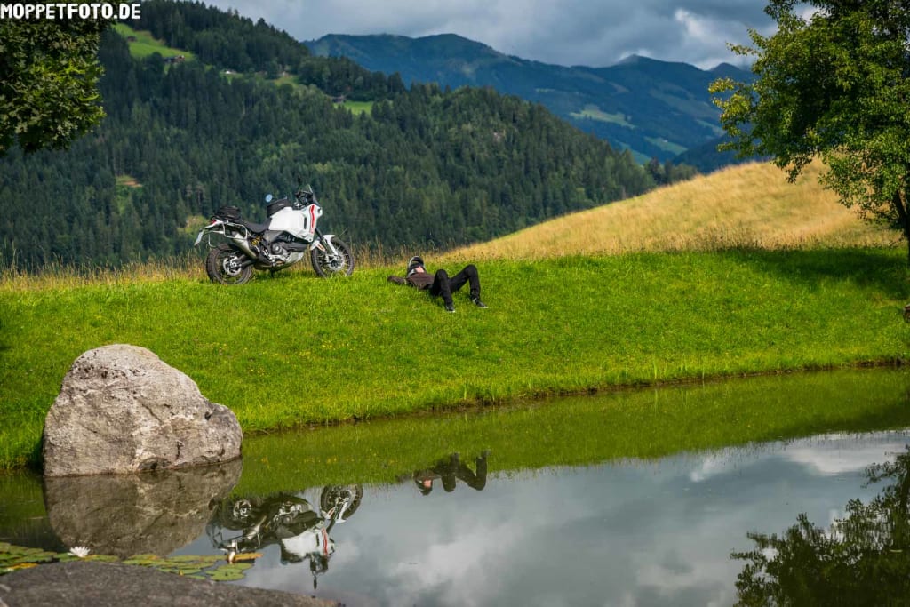Person liegt auf grüner Wiese mit Motorrad neben Teich und Bergkulisse