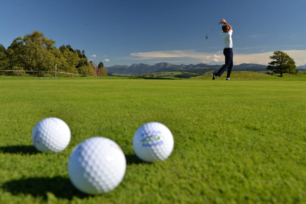 Golfschnupperkurs Golfspieler beim Abschlag auf grünem Golfplatz mit Bergen im Hintergrund