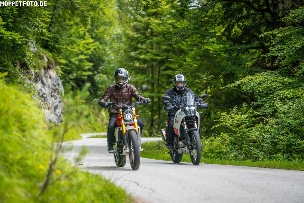Zwei Motorradfahrer fahren auf einer Straße durch einen grünen Wald