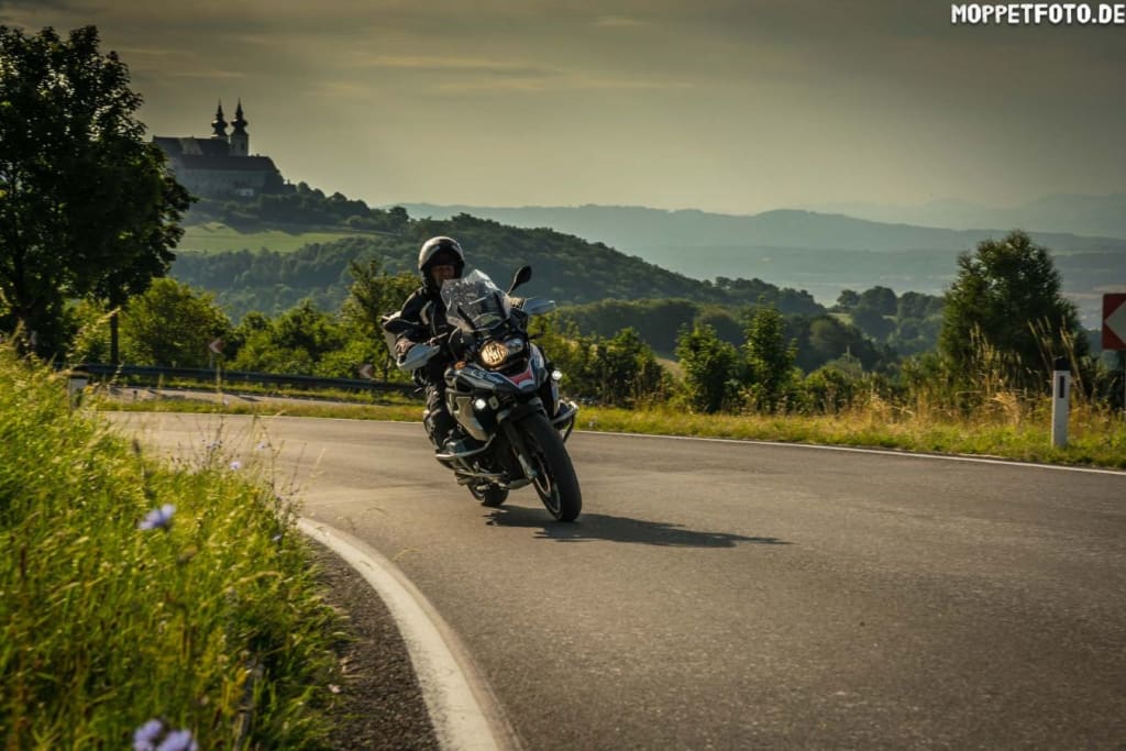 Motorradfahrer auf einer kurvigen Landstraße mit Hügeln und Schloss im Hintergrund