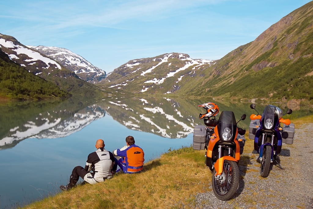 Zwei Motorradfahrer sitzen an einem See mit Bergspiegelung