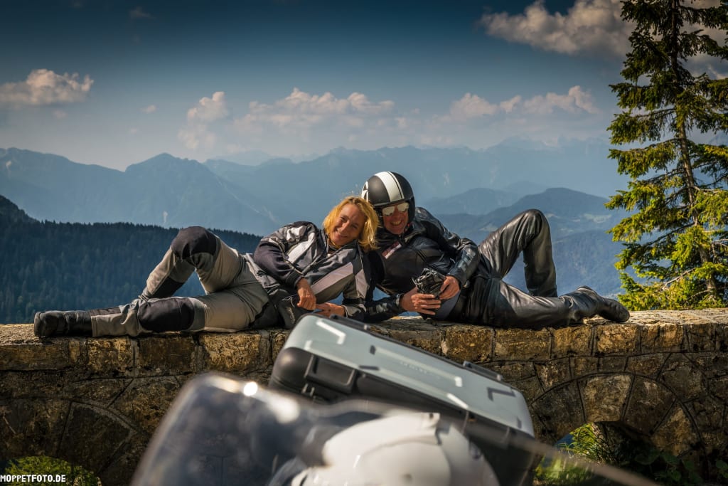 Zwei Motorradfahrer lehnen entspannt auf einer Steinmauer mit Berglandschaft im Hintergrund