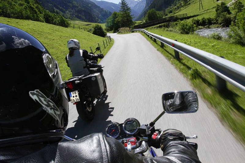 Zwei Motorradfahrer fahren auf einer kurvenreichen Bergstraße durch grüne Landschaft