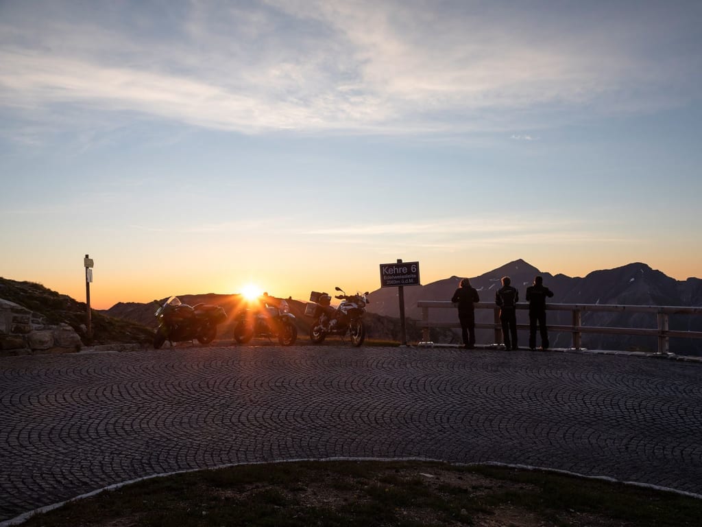Sternfahrt Motorrad-Special ab € 598,- pro Person Motorräder und Menschen bei Sonnenaufgang an einem Bergpass mit Schild Kehre 6