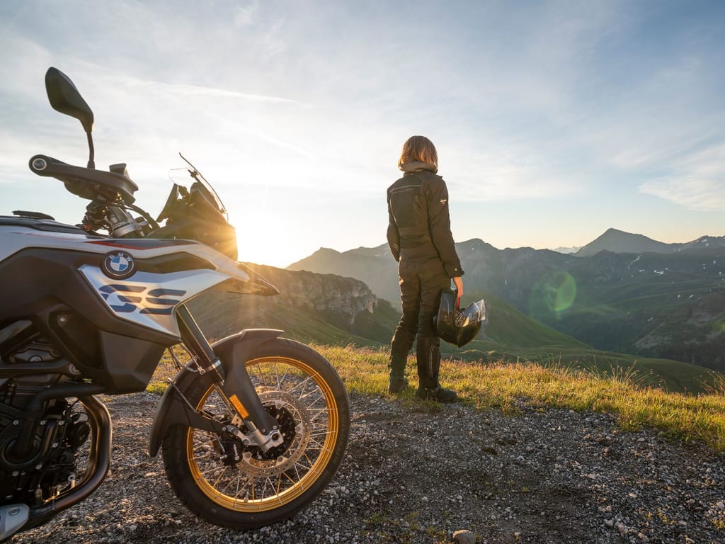 Auf die Berge - fertig - los ! ab € 398,- pro Person Motorradfahrer steht mit Helm auf bergigem Aussichtspunkt bei Sonnenuntergang